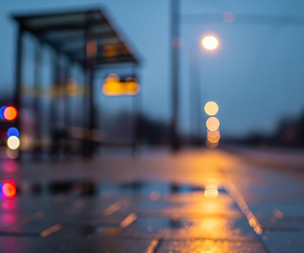 Rozmyty obraz przedstawia nocny widok ulicy po deszczu. Na pierwszym planie, w dolnej części, widoczne są kałuże i mokra nawierzchnia, odbijające jaskrawe, kolorowe światła. Po lewej stronie znajdują się rozmyte czerwone i niebieskie światła, przypominające lampy pojazdu, oraz ich odbicia w kałużach. W centrum kadru dominuje jasne, pomarańczowe światło latarni ulicznej, której blask rozlewa się po mokrej drodze, tworząc smugę odblasków. W tle, po lewej stronie, zarysowana jest konstrukcja przystanku autobusowego z niewyraźnym, żółtym wyświetlaczem. Całe tło tonie w odcieniach ciemnego błękitu i szarości, z kilkoma niewyraźnymi punktami świetlnymi, sugerującymi dalsze oświetlenie ulicy.