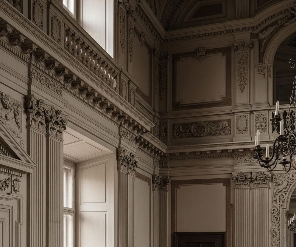 Wnętrze obszernej sali ukazuje bogato zdobione ściany w neutralnych odcieniach beżu i brązu, z wyraźnymi detalami sztukaterii. Po prawej stronie dominuje duży, żelazny żyrandol z licznymi ramionami i białymi świecami, zawieszony na tle wysokiej, arkadowej niszy, w której odbija się kolejny, mniejszy żyrandol. Lewa strona zdjęcia przedstawia fragment ściany z drzwiami ozdobionymi motywami roślinnymi i geometrycznymi, a dalej widać okno z subtelnie wpadającym światłem, które rozjaśnia górne partie balustrady, tworząc kontrast z ciemniejszymi elementami wnętrza.