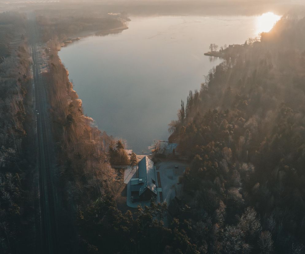 Widok z lotu ptaka na obiekt Boho Pogoria w Dąbrowie Górniczej, otoczony lasem i jeziorem o wschodzie słońca. Na zdjęciu widać budynek z długim spadzistym dachem, zlokalizowany blisko linii brzegowej, co podkreśla jego integrację z naturą, o czym można przeczytać na Architektura Murator Plus.