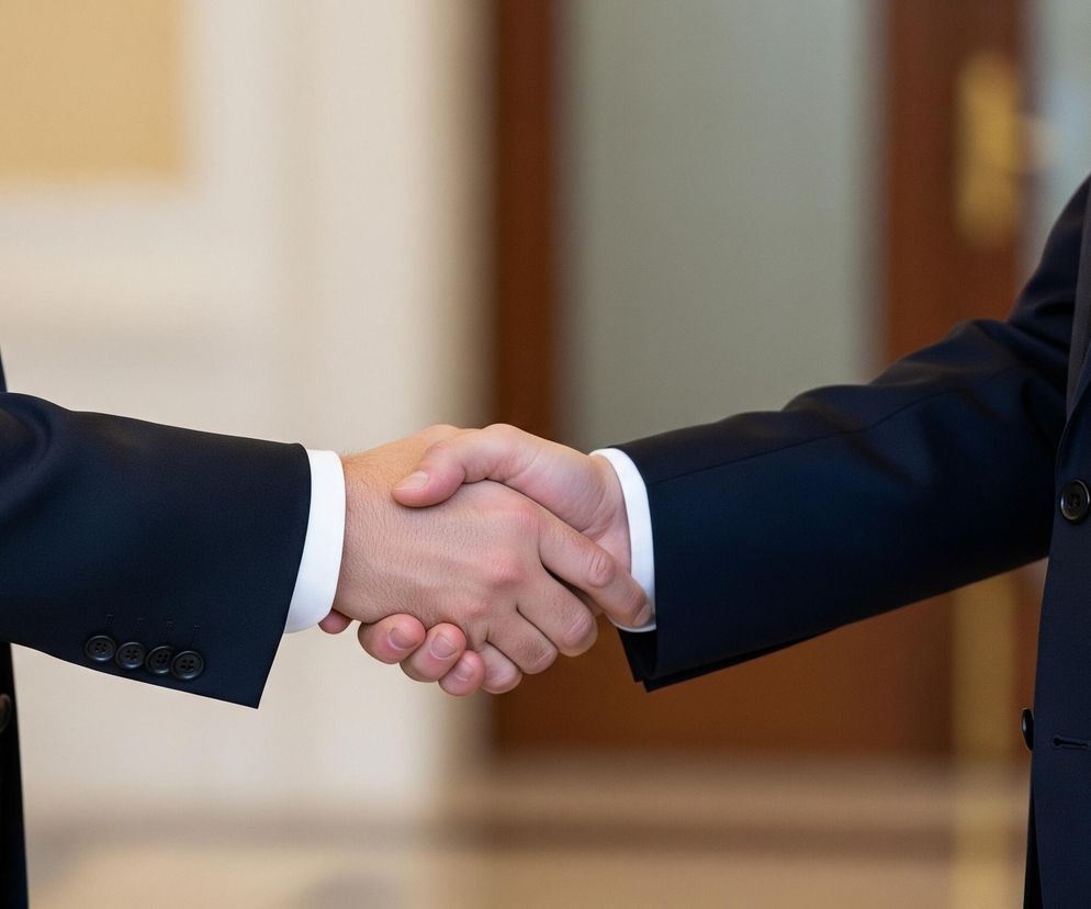 Dwie osoby w garniturach, widoczne od torsu w górę, ściskają sobie dłonie na pierwszym planie, dominując centralną część kadru. Lewa osoba ma na sobie ciemny garnitur z białą koszulą i fragmentem czerwonego krawata, natomiast prawa również ubrana jest w ciemny garnitur z białą koszulą i widocznym kawałkiem niebieskiego krawata. Tła stanowią rozmyte, beżowo-brązowe powierzchnie, sugerujące wnętrze.