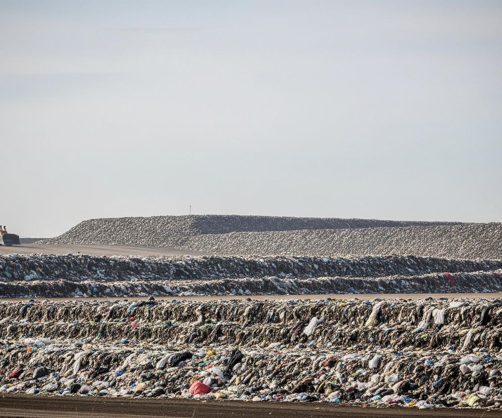 Rozległy krajobraz z widocznym wielkim składowiskiem odpadów rozciąga się pod jasnym, częściowo zachmurzonym niebem. W dolnej części zdjęcia, na pierwszym planie i w środkowej części, widać trzy poziomy ułożonych warstwowo, zbitych śmieci, składających się z różnobarwnych kawałków plastiku, tkanin i innych odpadów. Na horyzoncie, na szczycie dużego nasypu śmieci, widać dwie żółte koparki spychające kolejne odpady; jedna znajduje się po lewej stronie obrazu, druga po prawej.