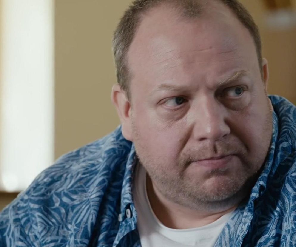 Zamyślony Bartłomiej Nowosielski jako Tadeusz z M jak miłość, ubrany w niebieską koszulę z białym wzorem, spogląda w bok z lekkim niepokojem. Ta scena z odcinka 1905 ukazuje Tadeusza podczas niezręcznej wizyty u lekarki Alicji, spowodowanej nietypową pomyłką z lakierem do włosów. To wydarzenie z pewnością zainteresuje czytelników portalu Super Seriale.