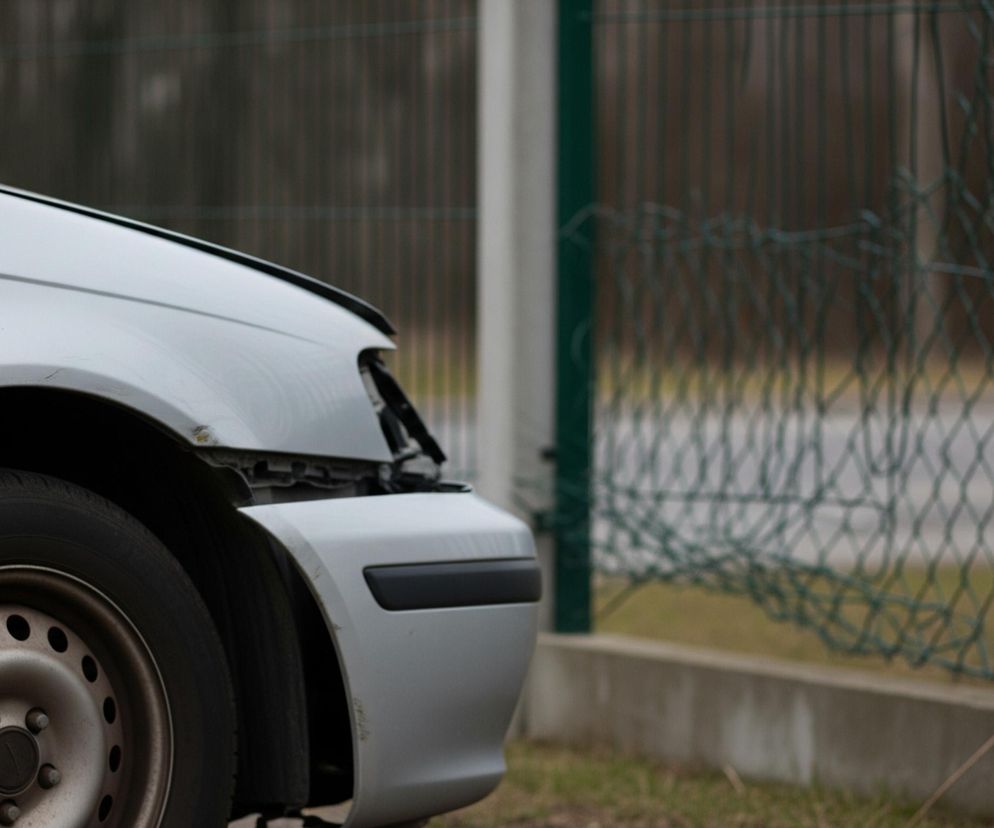 Srebrny samochód, ukazany w prawym dolnym rogu, stoi na nierównym, brudnym podłożu. Lewe przednie koło pojazdu, wraz z oponą i stalową felgą, jest wyraźnie widoczne, a na srebrnym błotniku i masce widać liczne rysy i zabrudzenia. Samochód ma uszkodzony zderzak i brakuje mu prawego przedniego reflektora, odsłaniając ciemne wnętrze komory silnika. W tle rozciąga się zielone ogrodzenie z siatki, lekko zdeformowane, a za nim niewyraźny krajobraz.