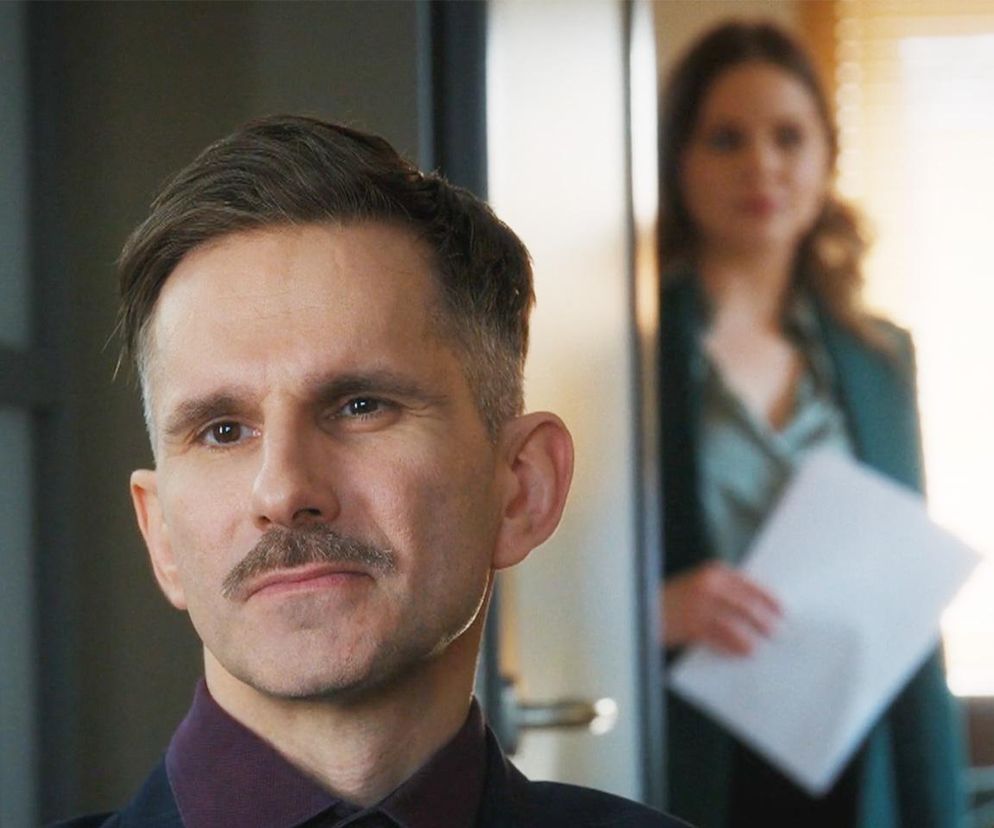 Marcin Bosak jako Kamil Gryc z wąsem, patrzy w lewo, a w tle rozmyta Melania Grzesiewicz jako Anita Laskowska z dokumentami, symbolizujące tajemnicę Gryca. Więcej o ich losach w M jak miłość na Super Seriale.