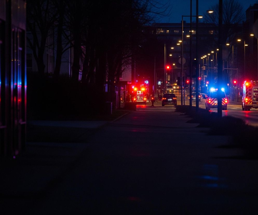 Ulica miejska o zmierzchu lub w nocy, z widocznymi światłami awaryjnymi pojazdów uprzywilejowanych. Po lewej stronie widać odbicia czerwonych i niebieskich świateł na pionowych, szklanych powierzchniach budynku. Droga prowadzi prosto w głąb kadru, oświetlona szeregiem żółtych lamp ulicznych i czerwonymi światłami sygnalizacji. Na jezdni znajduje się kilka samochodów, w tym ciężarówka strażacka z włączonymi czerwonymi światłami awaryjnymi oraz pojazd z niebieskimi światłami uprzywilejowania.