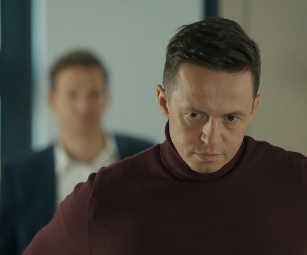 Piotr Ligienza jako Błażej w serialu Barwy szczęścia, w bordowym golfie z poważnym, spiskującym wyrazem twarzy, patrzący prosto w obiektyw. W tle, niewyraźna postać Michała Rolnickiego jako Łukasza. Ten kadr sugeruje intrygę i konflikt, o których więcej przeczytasz na portalu Super Seriale.