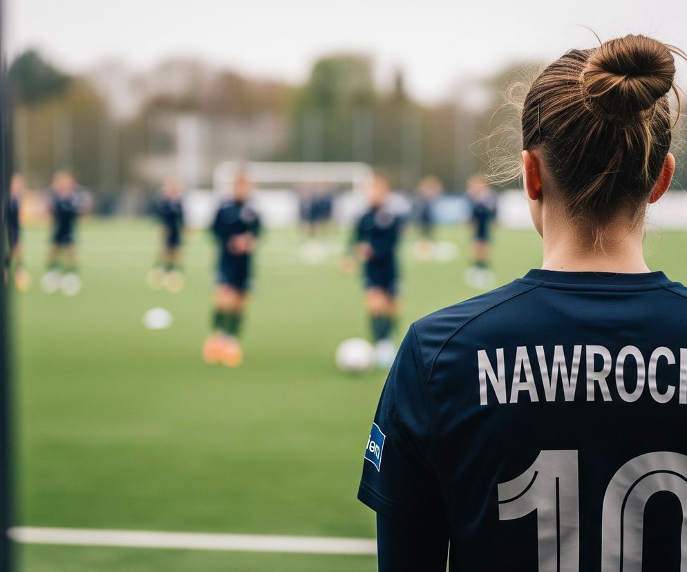 Kobieta z upiętymi włosami stoi tyłem do obserwatora, ubrana w granatową koszulkę sportową z nazwiskiem NAWROCKA i numerem 10 na plecach, patrząca na boisko. Na boisku, pokrytym zieloną sztuczną trawą, w tle widać rozmyte sylwetki innych osób w ciemnych strojach sportowych, a także fragmenty białych linii i pachołków treningowych. Po lewej stronie zdjęcia widoczny jest pionowy, ciemny element architektoniczny, który częściowo zasłania widok na boisko.