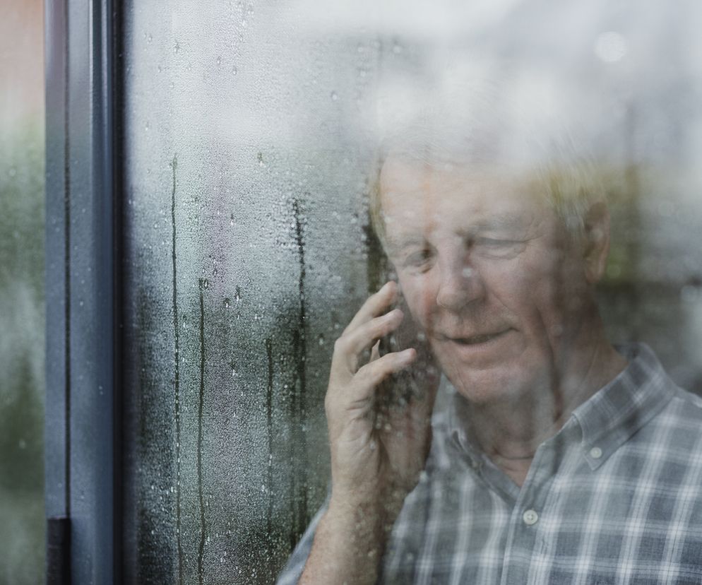 Samotny starszy mężczyzna rozmawia przez telefon, patrząc przez zaparowane okno. Scena odzwierciedla poczucie izolacji i potrzebę kontaktu, co jest kluczowe dla zdrowia psychicznego seniorów. Dowiedz się więcej o wsparciu dla samotnych seniorów na Poradnik Zdrowie.