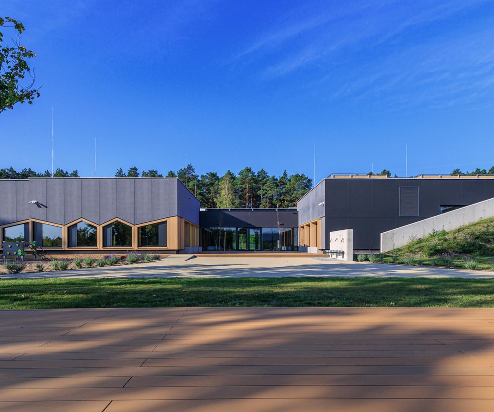 Nowoczesne Przedszkole w Bydgoskim Parku Przemysłowo-Technologicznym pracowni xystudio, charakteryzujące się prostą bryłą, szarymi elewacjami z elementami drewnianymi oraz oknami w kształcie domków. Widoczny teren zielony z górkami i placem zabaw, idealne miejsce do poznania nowości w Architektura Murator Plus.