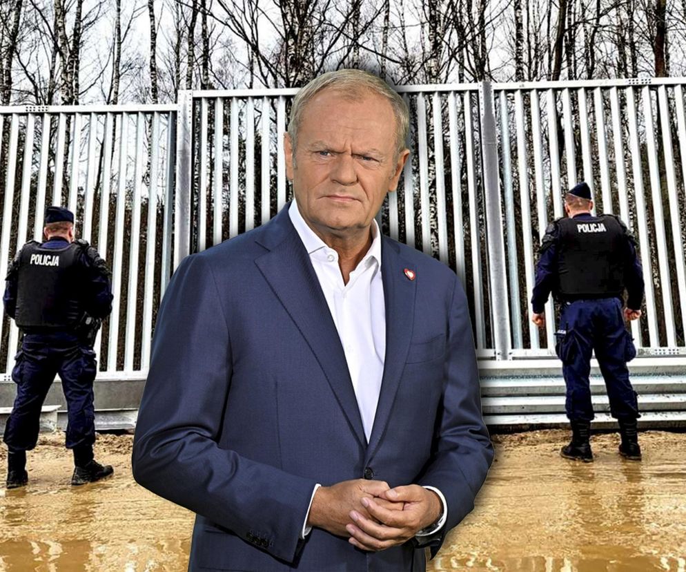 Donald Tusk w granatowym garniturze i białej koszuli, z zamyślonym wyrazem twarzy, na tle stalowego ogrodzenia granicznego i dwóch policjantów. Fotografia symbolizuje decyzję premiera o otwarciu granicy z Białorusią. Więcej o tej decyzji przeczytasz na Super Biznes.