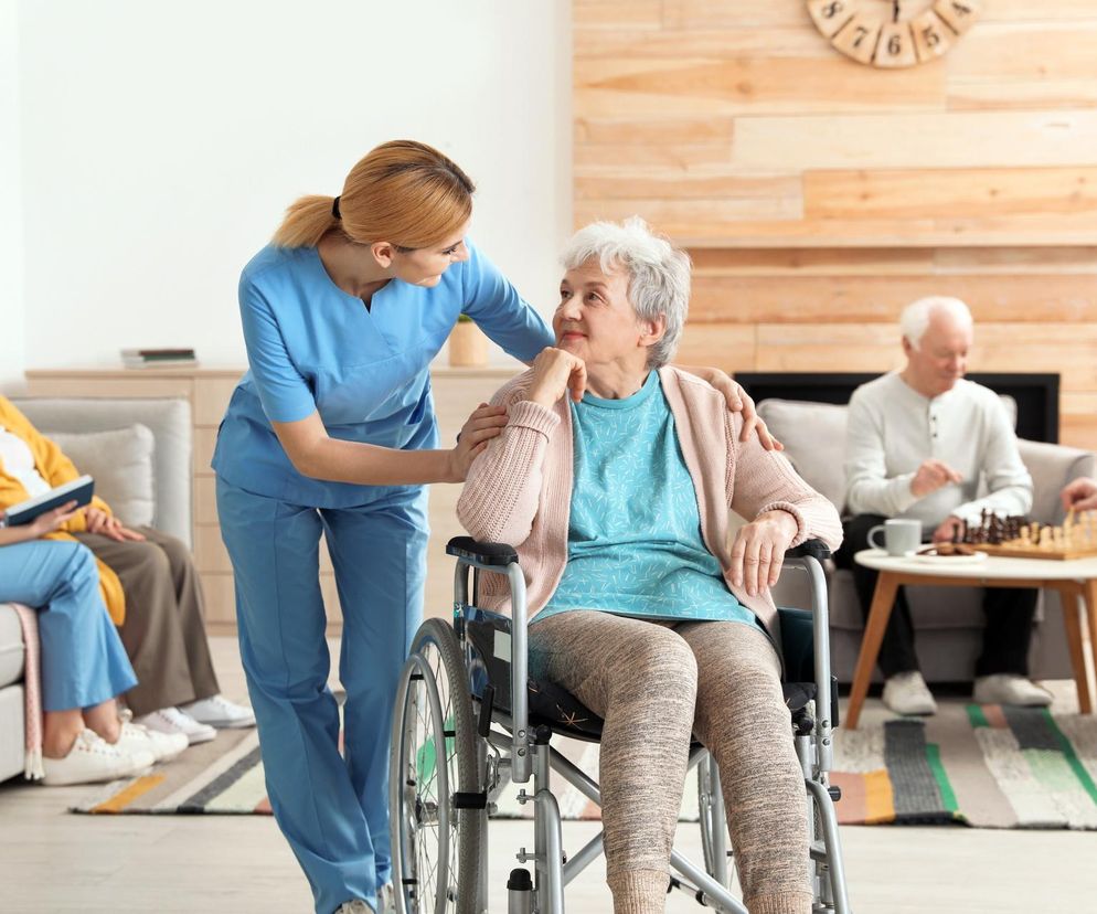 Pielęgniarka w niebieskim uniformie wspiera starszą kobietę na wózku inwalidzkim w domu seniora. W tle inni pensjonariusze. Artykuł na Super Biznes informuje o szokujących kosztach pobytu w domach opieki w Niemczech.