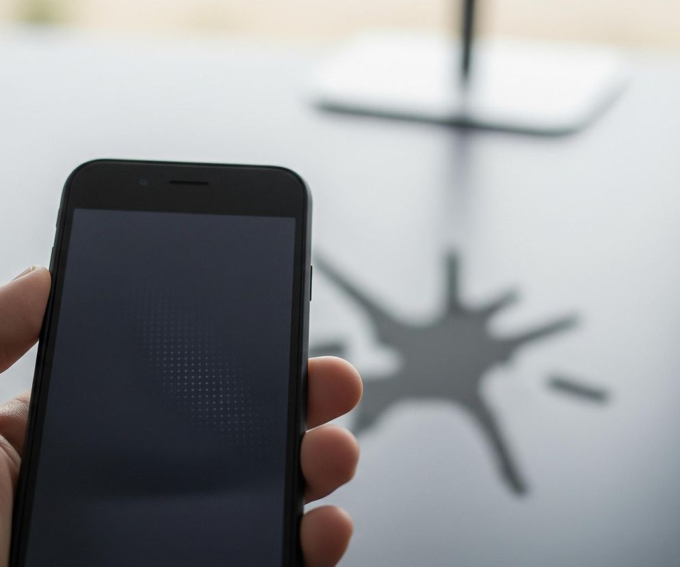 Lewa dłoń trzyma czarny smartfon, którego ekran jest wyłączony i ma ciemnoszare zabarwienie z widocznym wzorem kropek. Górna część telefonu ukazuje niewielki otwór i podłużną szczelinę, a także błyszczącą ramkę. W tle, na jasnoszarym blacie, rozmyty jest ciemny cień o nieregularnym, gwiazdkowatym kształcie z kilkoma ostrymi ramionami.