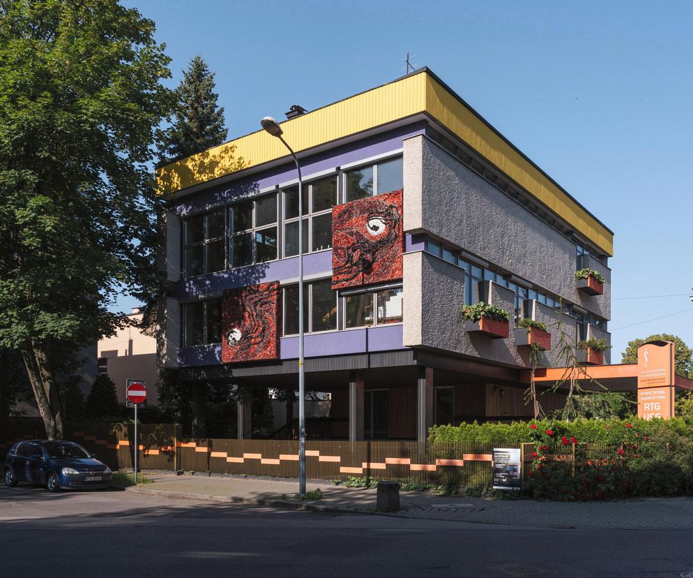 Modernistyczny Dom Książków w Tarnowie z charakterystyczną fioletową elewacją, szarymi fragmentami i żółtym dachem, ozdobiony mozaikami. Na pierwszym planie ulica i zaparkowany samochód. Więcej o architekturze powojennej znajdziesz na Architektura Murator Plus.