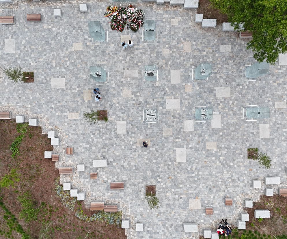 Widok z góry na skwer z Pomnikiem Żołnierzy Niezłomnych we Wrocławiu, z granitową kostką, ławkami, kamiennymi blokami i zielenią. W centralnej części widać szklane postacie żołnierzy oraz ułożone wieńce, co nawiązuje do pamięci i refleksji. O projekcie przeczytasz na Architektura Murator Plus.