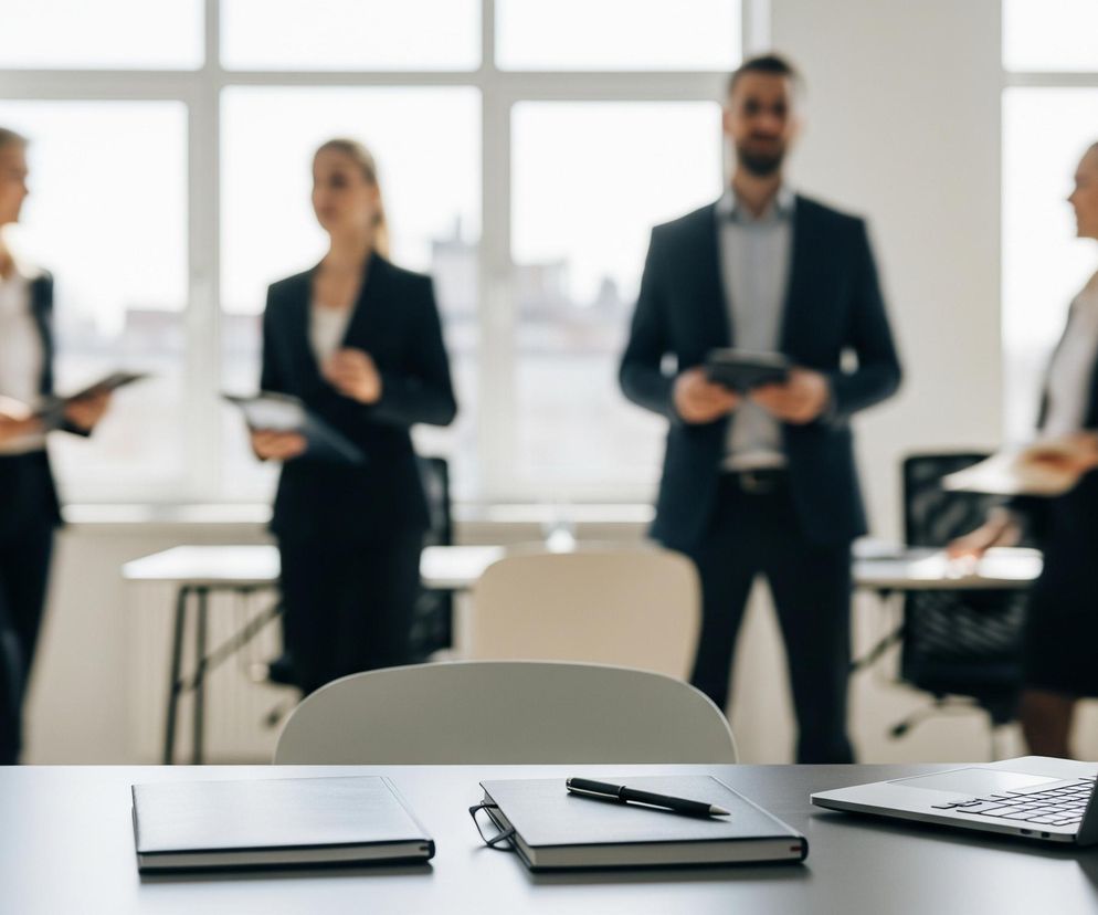 Na pierwszym planie, na ciemnoszarym blacie biurka, leży otwarty laptop w kolorze srebrnym, zwrócony ekranem w prawo, z widoczną klawiaturą. Obok niego, bliżej środka, znajduje się ciemny notatnik z czarnym długopisem ułożonym na nim. Po lewej stronie leży kolejny ciemny, zamknięty notatnik. Za biurkiem widać jasne krzesło.

W tle, ostrość jest rozmyta, ale widać cztery sylwetki osób stojących. Dwie kobiety i mężczyzna, wszyscy ubrani w formalne stroje, z czego dwie kobiety i mężczyzna w centrum trzymają coś w rękach, przypominające tablety lub notesy. Cała scena rozgrywa się w jasnym pomieszczeniu z dużymi oknami w tle, przez które wpada dzienne światło.