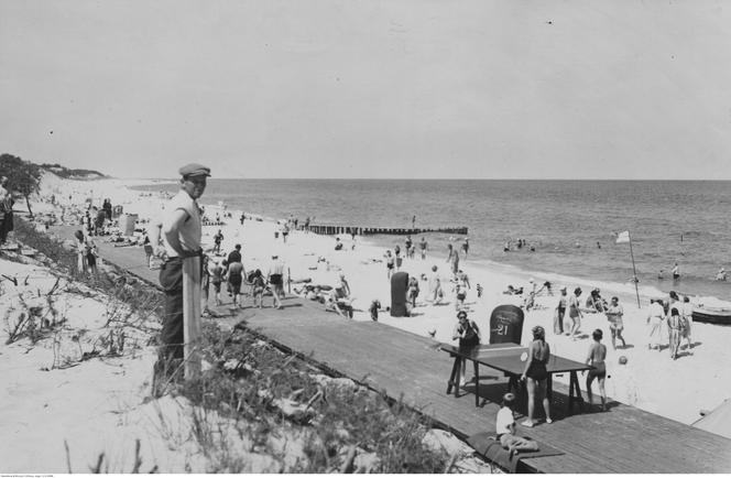 Plażowicze podczas wypoczynku. Widoczny drewniany deptak, wiklinowe kosze plażowe oraz stół do ping-ponga. Zdjęcie zrobiono w okresie pomiędzy 1918-1937 roku w Juracie 