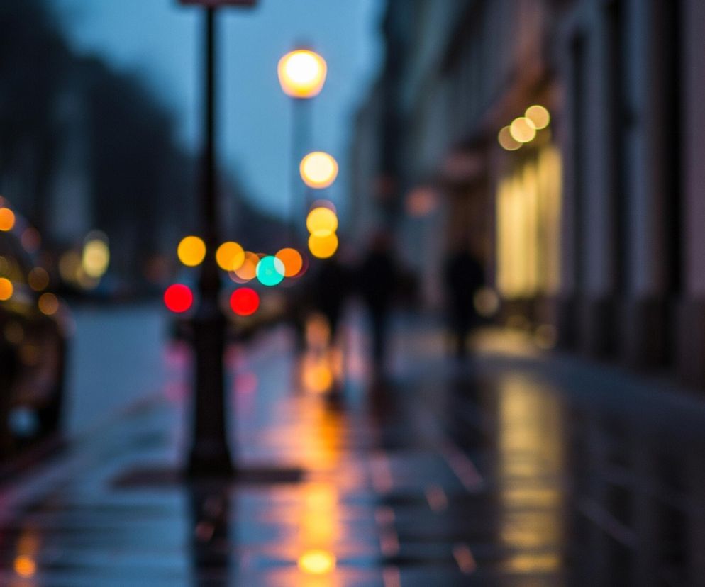 Zamazana scena miejska ukazuje czarny samochód zaparkowany po lewej stronie kadru, z jego czerwonymi światłami odbijającymi się w mokrym asfalcie. Centralnie, widoczny jest czarny słup latarni ulicznej, a wokół niego i w oddali rozmyte, kuliste światła miejskie w odcieniach pomarańczowego, czerwonego i turkusowego. Prawo od słupa, rozmyte sylwetki ludzi spacerują po chodniku, na którym również widoczne są odbicia świateł. Całość utrzymana jest w ciemnych, chłodnych barwach, z ciepłymi akcentami świetlnymi.