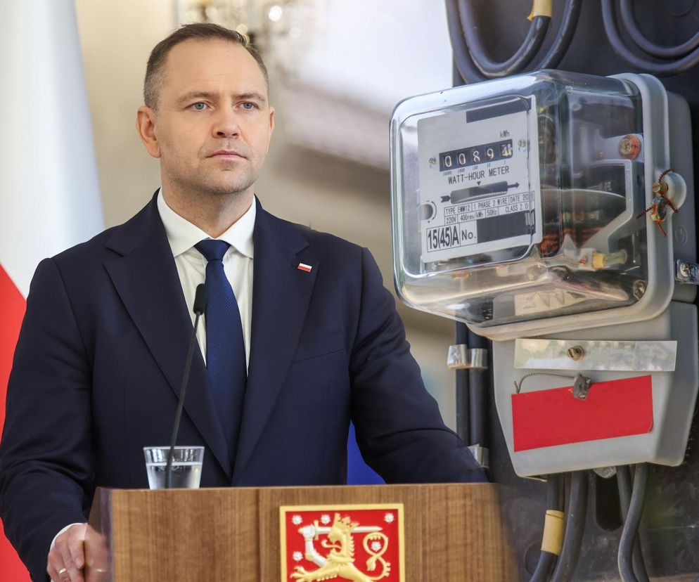 Karol Nawrocki, prezes Instytutu Pamięci Narodowej, w ciemnym garniturze i krawacie, stojący za mównicą z polską flagą w tle, obok niego licznik prądu symbolizujący mrożenie cen energii elektrycznej. Temat ważny dla czytelników Super Biznes.