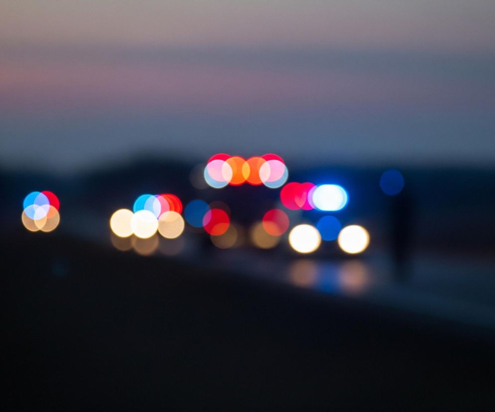 Rozmyte światła pojazdów na drodze o zmierzchu lub w nocy, z efektem bokeh. W centrum widać skupisko okrągłych, jaskrawych świateł w kolorach czerwonym, niebieskim i żółto-białym, sugerujących obecność samochodów, w tym prawdopodobnie pojazdu uprzywilejowanego. Dolna część kadru jest ciemna, symbolizując drogę, a górna część to rozmyte, ciemniejsze niebo z delikatnymi odcieniami fioletu i szarości.