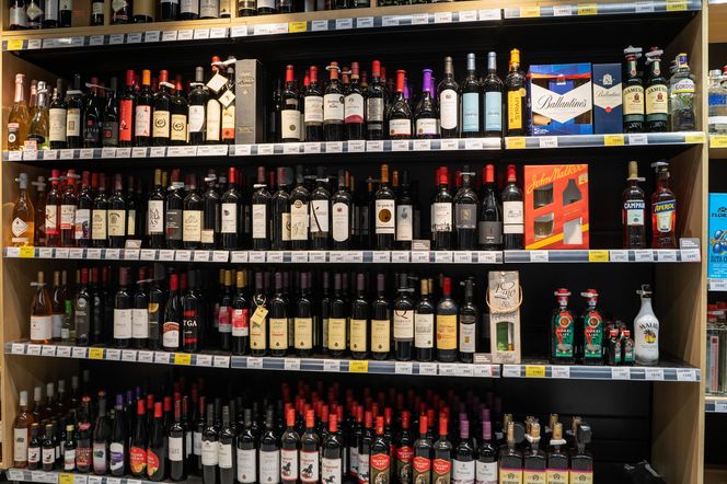 Duży regał w sklepie, na którym widać liczne butelki z różnymi rodzajami alkoholu, w tym wina, whisky, wódki oraz likiery, ułożone w rzędach, z widocznymi cenami. Ta wizualizacja dostępności alkoholu podkreśla apel lekarzy o nocną prohibicję, o czym przeczytasz na Poradnik Zdrowie.