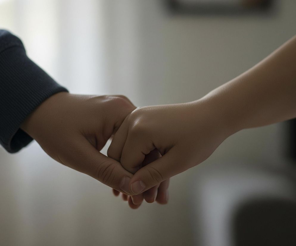 Dwie ręce, należące do różnych osób, są splecione, tworząc ciasny uścisk. Lewa ręka, z ciemniejszą skórą, ubrana jest w granatowy rękaw z prążkowanym ściągaczem na nadgarstku. Prawa ręka, o jaśniejszej karnacji, jest naga i ma delikatnie zgięte palce, a jej przedramię rozciąga się w prawą stronę obrazu. Tło jest jasne i rozmyte, z widocznymi odcieniami szarości i bieli, sugerującymi wnętrze pomieszczenia z naturalnym światłem.