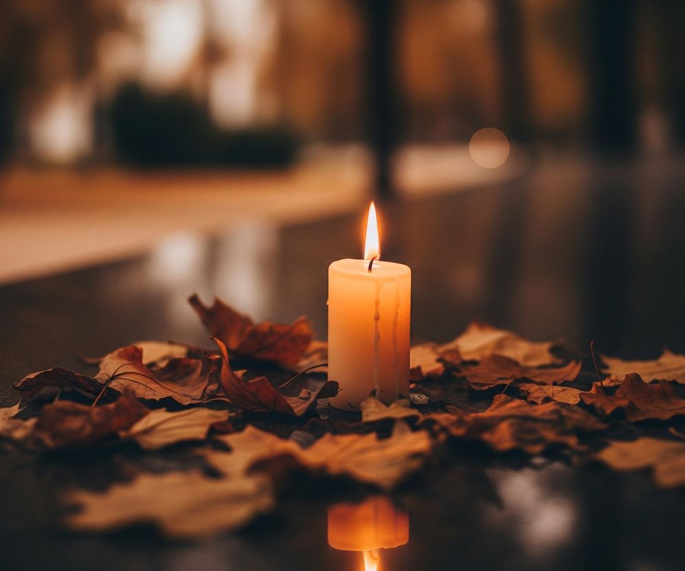 Na pierwszym planie widoczna jest zapalona świeca o barwie kremowo-pomarańczowej, otoczona suchymi, jesiennymi liśćmi w odcieniach brązu i pomarańczu. Świeca stoi na ciemnej, gładkiej powierzchni, która odbija jej płomień i część wosku, tworząc lustrzane odbicie. Tło jest rozmyte, utrzymane w ciepłych, ciemnych tonach, z kilkoma jaśniejszymi punktami świetlnymi, co sugeruje scenerię wieczorną lub nocną.