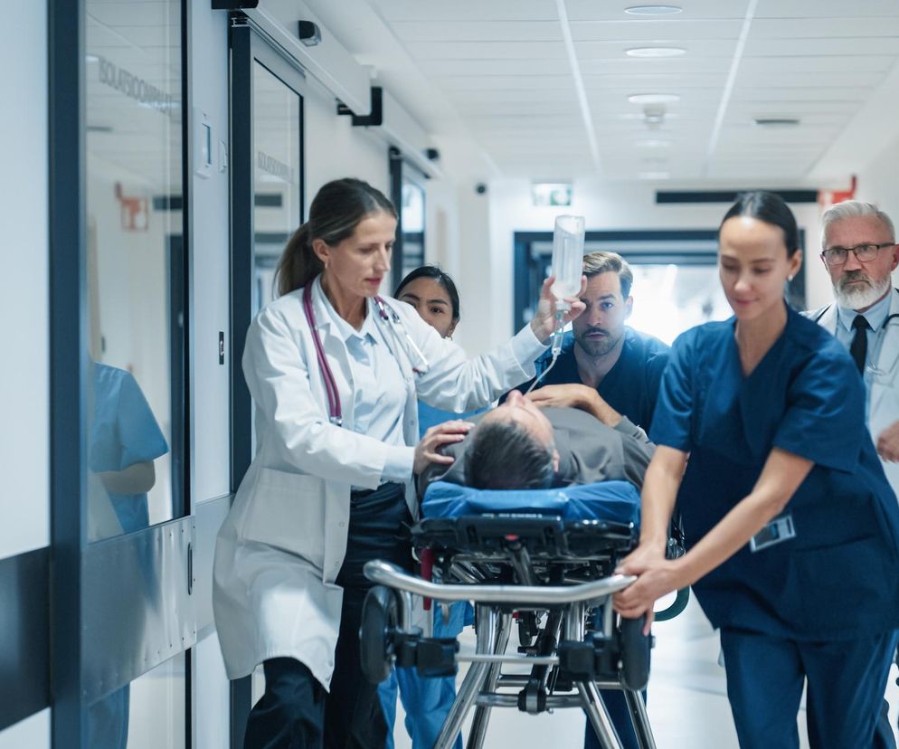 Zespół medyczny w biegu transportuje pacjenta na noszach przez szpitalny korytarz, co symbolizuje pilną pomoc w obliczu zagrożeń zdrowotnych. Artykuł na Poradnik Zdrowie przestrzega przed mięsożerną bakterią Vibrio vulnificus.