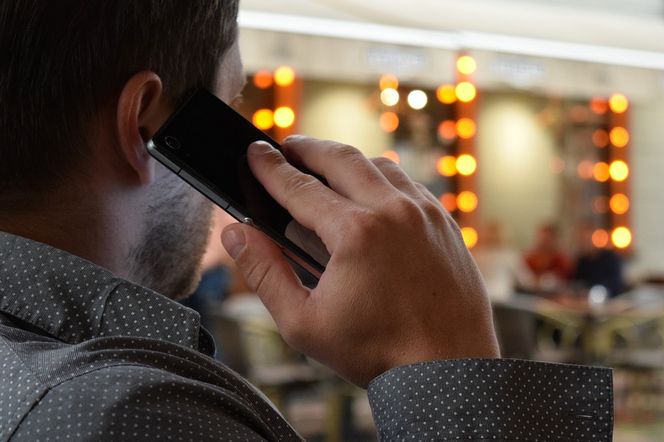 Mężczyzna w szarej koszuli w kropki rozmawia przez telefon, trzymając smartfon przy uchu, jego palec wskazujący dotyka ekranu. Tło jest rozmazane, widać jasne, pomarańczowe światła i zarysy osób. Zdjęcie ilustruje problemy z telekomunikacją w kontekście awarii Plus, o czym można przeczytać na Super Biznes.