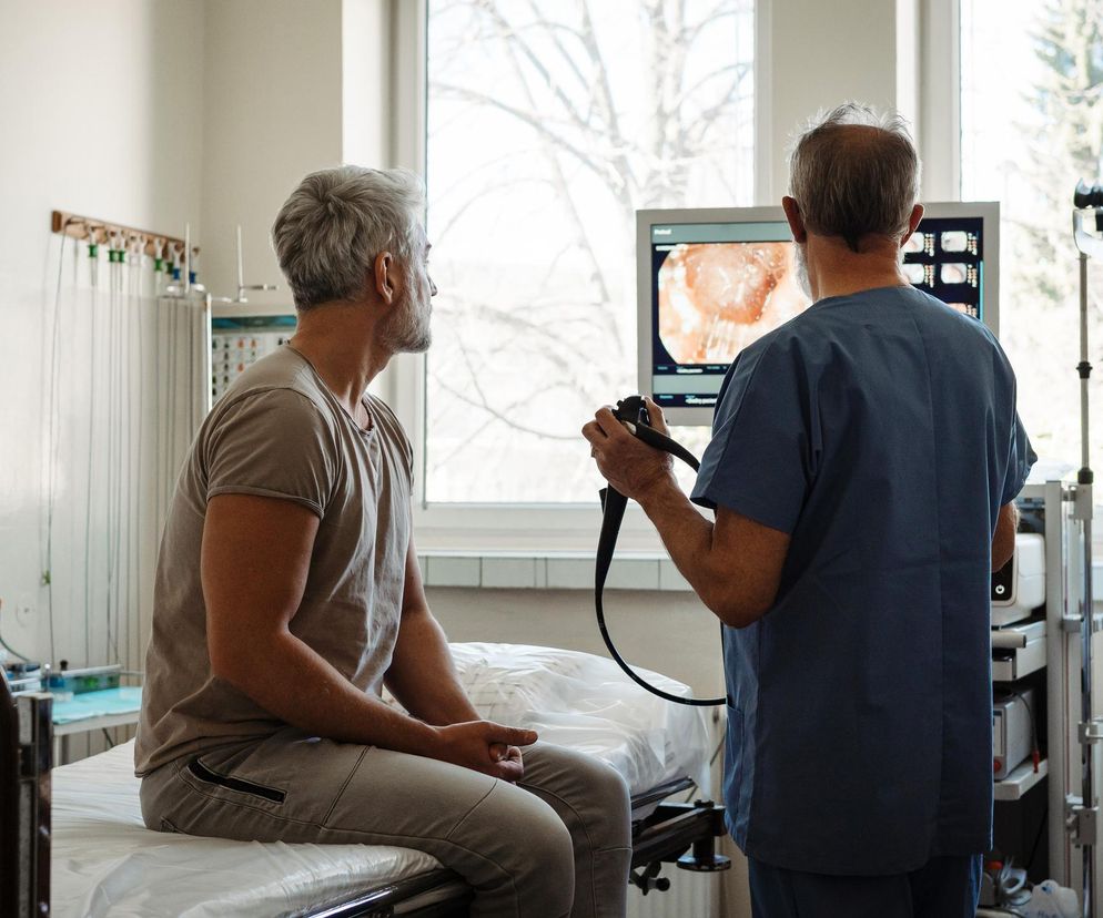 Pacjent siedzący na leżance w gabinecie lekarskim, obserwujący monitor z obrazem z kolonoskopii, którą trzyma lekarz. Scena podkreśla znaczenie badań przesiewowych w profilaktyce raka jelita grubego, o czym przeczytasz więcej na Poradnik Zdrowie.