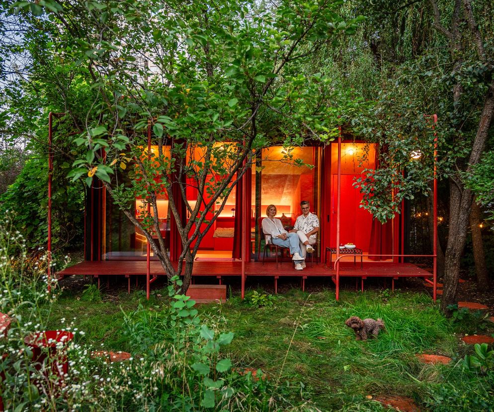 Czerwony, nowoczesny dom letniskowy ROD. Dom koloru autorstwa Noke Architects, zlokalizowany w otoczeniu zieleni, z widocznymi właścicielami, Magdą Grabowską-Wacławek i Grzegorzem Wacławkiem, siedzącymi na tarasie. O projekcie przeczytasz na Architektura Murator Plus.