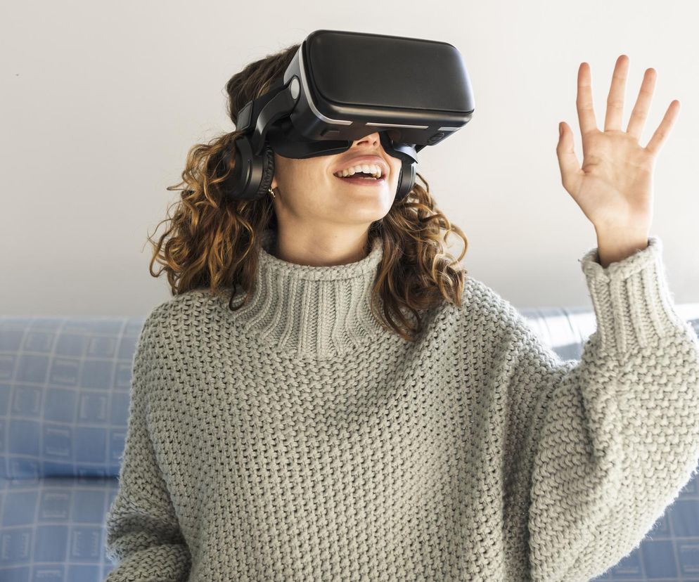 Kobieta z kręconymi włosami w szarym swetrze siedzi na kanapie, używając gogli VR z szerokim uśmiechem, podnosząc prawą dłoń. Odczuwa relaks, co ilustruje artykuł o tym, jak jesień nie musi być przygnębiająca i o sposobach redukcji stresu, o czym przeczytasz na Poradnik Zdrowie.