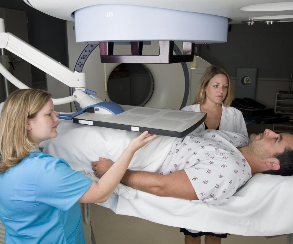 Mężczyzna leżący na stole do radioterapii, otoczony przez dwie lekarki. Jedna z nich kładzie na jego klatce piersiowej płytkę, druga obserwuje. Sprzęt medyczny widoczny w tle. Więcej o pielęgnacji skóry podczas radioterapii przeczytasz na Poradnik Zdrowie.