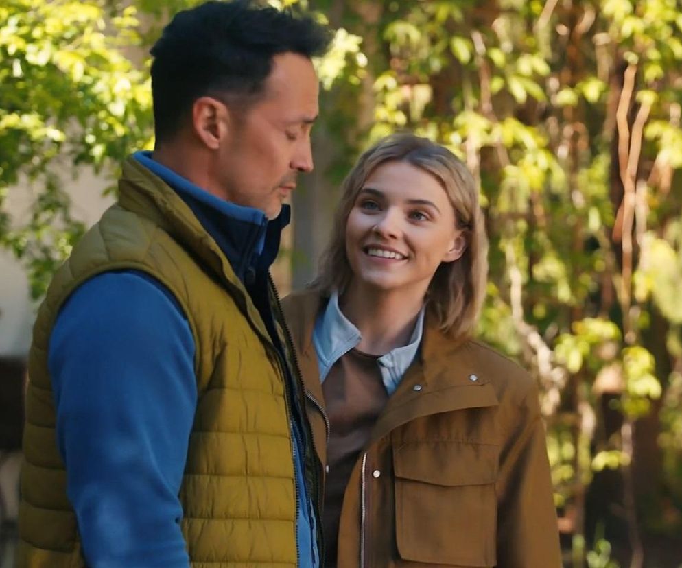 Mariusz (Rafał Cieszyński) w żółtej kamizelce i niebieskiej bluzie z długim rękawem oraz Janka (Oliwia Drożdżyk) w brązowej kurtce i jasnej koszuli, patrzą na siebie z uśmiechem w zielonym otoczeniu. Zdjęcie przedstawia scenę z serialu Barwy szczęścia, o którym więcej przeczytasz na portalu Super Seriale.