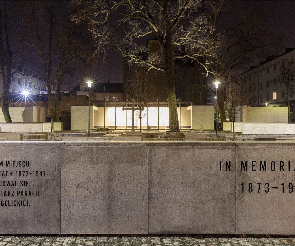 Oświetlony plac, upamiętniający dawny cmentarz ewangelicki, z betonowymi płytami, na których widnieją napisy W TYM MIEJSCU W LATACH 1873-1947 ZNAJDOWAŁ SIĘ CMENTARZ PARAFII EWANGELICKIEJ oraz IN MEMORIAM 1873-1947. W tle widoczne nowoczesne budynki węzła przesiadkowego w Olsztynie, autorstwa Architekci GADOMSCY. O tym i innych projektach przeczytasz na Architektura Murator Plus.