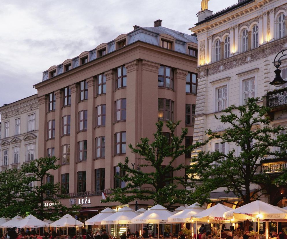 Dom Feniksa w Krakowie, modernistyczna kamienica z mansardowym dachem, otoczona zabytkowymi budynkami na Rynku Głównym. Przed budynkiem restauracyjny ogródek z białymi parasolami, tętniący życiem po zmierzchu. O historii Feniksa przeczytasz na Architektura Murator Plus.