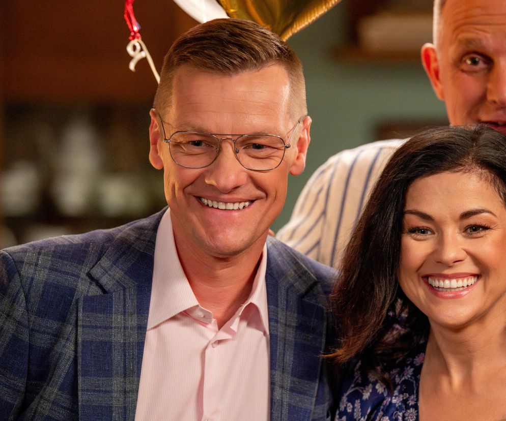 Piotrek i Kinga Zduńscy z M jak miłość uśmiechają się radośnie, stojąc obok siebie. Kinga ma na sobie granatową sukienkę w kwiaty. Uśmiechnięty Piotrek, w okularach i eleganckiej marynarce w kratę, z tyłu widać fragmenty innych osób. Więcej o ich losach przeczytasz na Super Seriale.