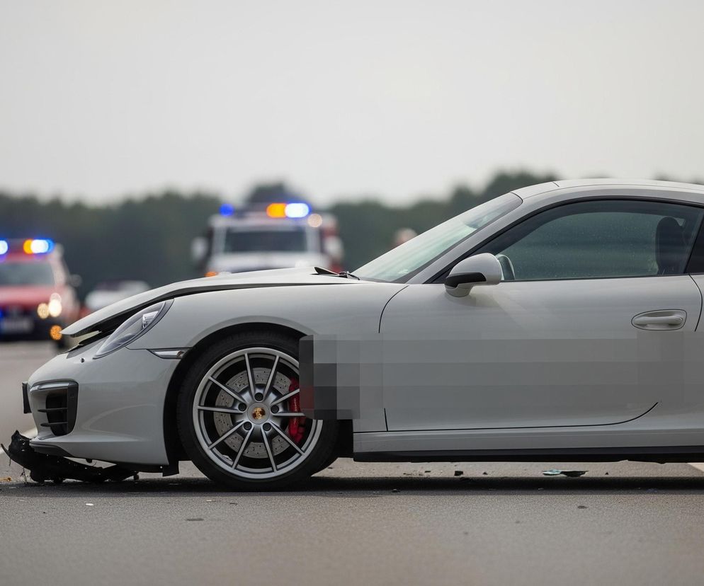 Biały samochód sportowy stoi na poboczu drogi asfaltowej, z przodem mocno uszkodzonym, a części zderzaka leżą na jezdni. Koła pojazdu są srebrne z czerwonymi zaciskami hamulcowymi, a na masce widać wgniecenia. W tle, na środkowym pasie drogi, widać dwa pojazdy uprzywilejowane z włączonymi niebieskimi i pomarańczowymi światłami sygnalizacyjnymi, jeden z nich to czerwony wóz strażacki, drugi biały pojazd prawdopodobnie medyczny lub policyjny. Horyzont zasłania pas drzew pod szarym, zachmurzonym niebem.