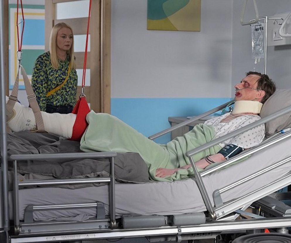 Fryderyk Graff leży pobity w szpitalnym łóżku, z opatrunkiem na nodze i kołnierzem ortopedycznym na szyi, podczas gdy Emilka patrzy na niego z zaniepokojeniem. Scena z serialu Pierwsza miłość ukazująca dramatyczne wydarzenia, o których przeczytasz więcej na Super Seriale.