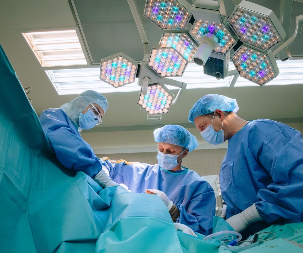 Zespół chirurgów podczas operacji w sali operacyjnej. Trzech lekarzy w niebieskich strojach chirurgicznych, maskach i czepkach wykonuje zabieg pod światłami operacyjnymi. Temat limitu zarobków dla lekarzy znajdziesz na Super Biznes.