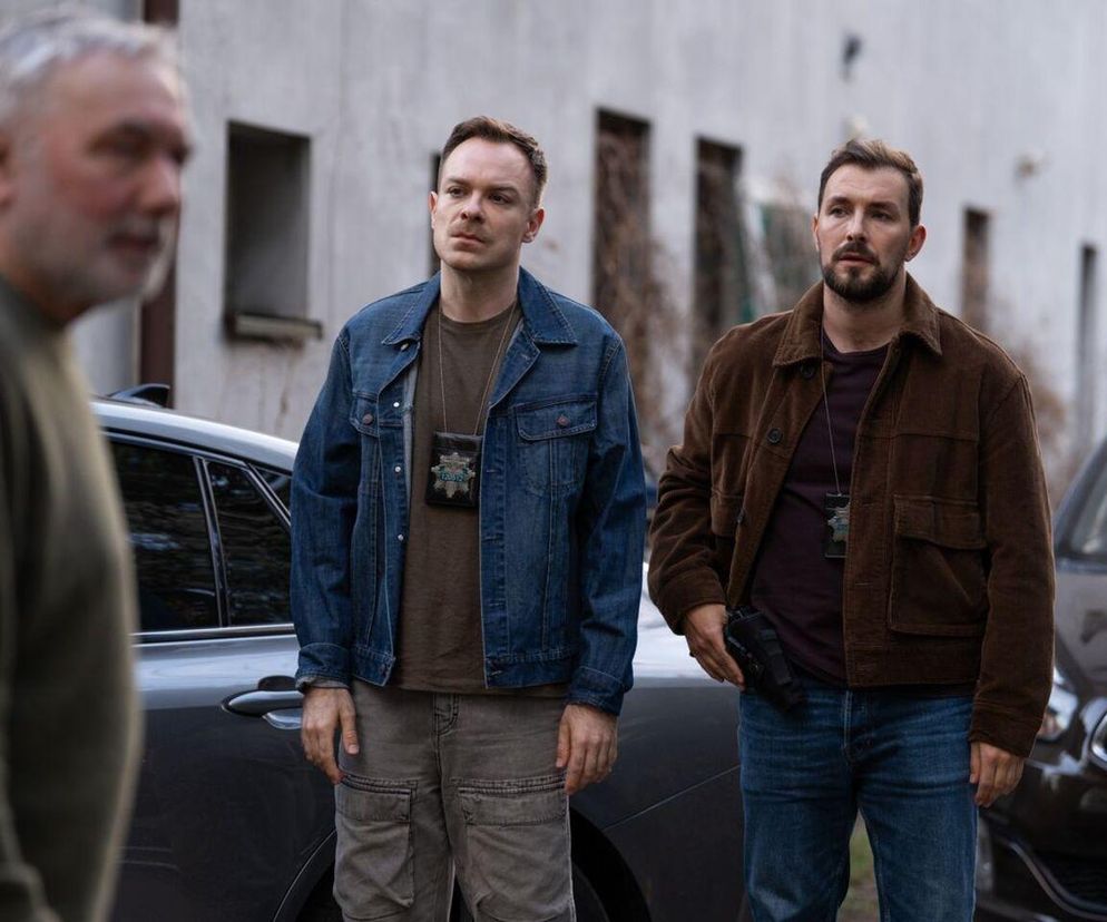 Komisarze Marcin Kodur i Majak zatrzymują Andrzeja, który stoi tyłem, rozmazany, w 3244 odcinku serialu Barwy szczęścia. Policjanci mają odznaki na szyi, pistolet w kaburze, a obok nich widać samochody. Więcej o ich losach na Super Seriale.