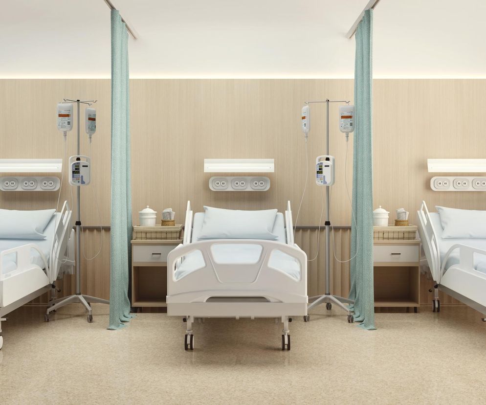 Trzy puste, białe łóżka szpitalne z niebieską pościelą, oddzielone turkusowymi zasłonami. Przy każdym łóżku stoją stoliki nocne oraz kroplówki z widocznymi urządzeniami medycznymi. Zdjęcie ilustruje problemy z przyjęciami pacjentów w szpitalach, o czym przeczytasz na Poradnik Zdrowie.