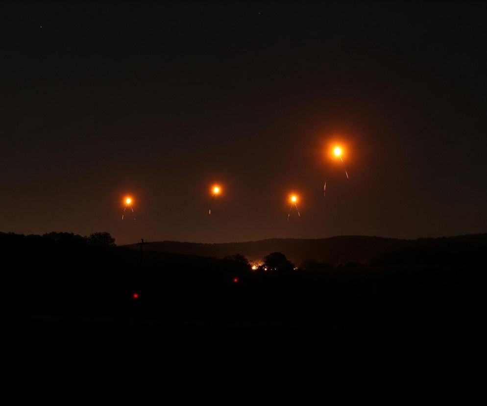 Cztery jasne, pomarańczowo-żółte punkty światła, wyglądające jak flary, unoszą się na ciemnym niebie nad horyzontem. Każda flara emituje smugę światła skierowaną w dół, sugerując opadanie. Poniżej rozciąga się ciemny krajobraz z zarysami wzgórz i drzew, a w oddali widać słabe światła naziemne oraz dwie małe czerwone kropki.