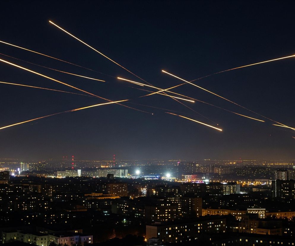 Nocne ujęcie miasta widzianego z góry, z licznymi liniami światła przecinającymi ciemne niebo. Na pierwszym planie i w środkowej części dominują ciemne dachy i fasady budynków, których okna świecą żółtym światłem, tworząc siatkę punktów świetlnych. W tle rozciąga się miejski krajobraz z rozproszonymi światłami, a horyzont jest lekko rozmazany. Powyżej miasta, na niemal czarnym niebie, widoczne są liczne jasne, żółtopomarańczowe smugi światła, biegnące w różnych kierunkach i tworzące efektowny wzór.