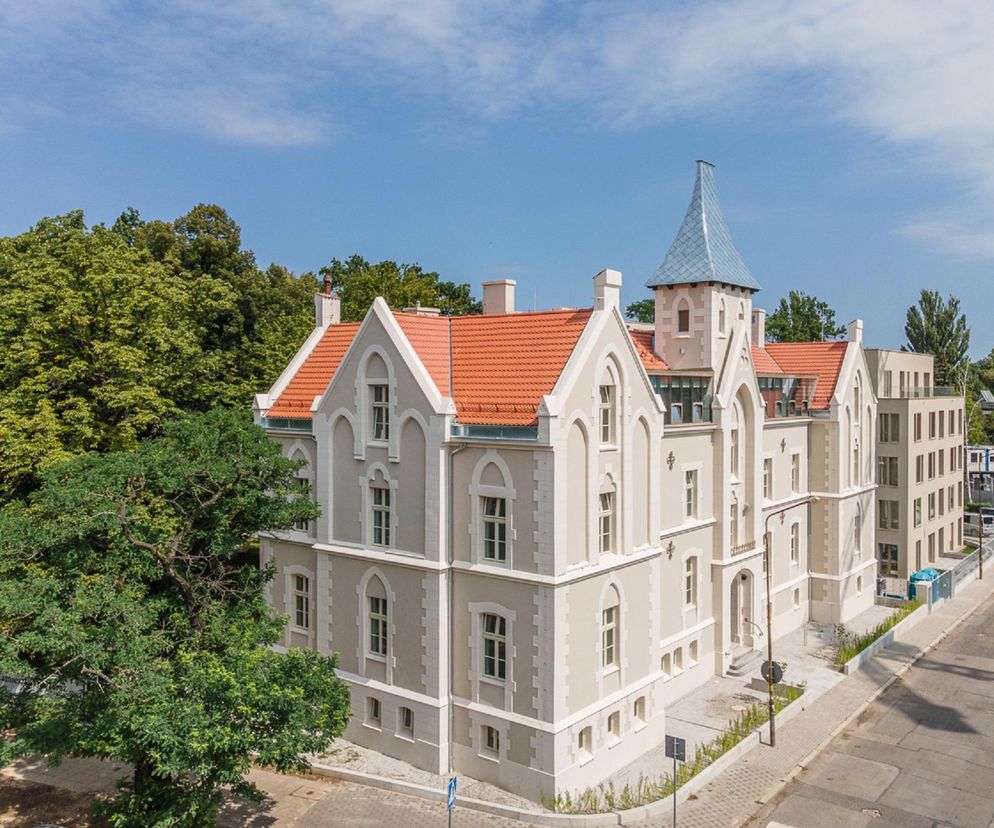 Neogotycki budynek z pomarańczowym dachem i charakterystyczną wieżyczką, sąsiadujący z nowoczesną, beżową zabudową apartamentową oraz zielonymi drzewami. Projekt rewitalizacji Ogrodów Graua we Wrocławiu, zgłoszony przez Maćków Pracownia Projektowa do konkursu Życie w Architekturze 2025, o którym przeczytasz więcej na Architektura Murator Plus.