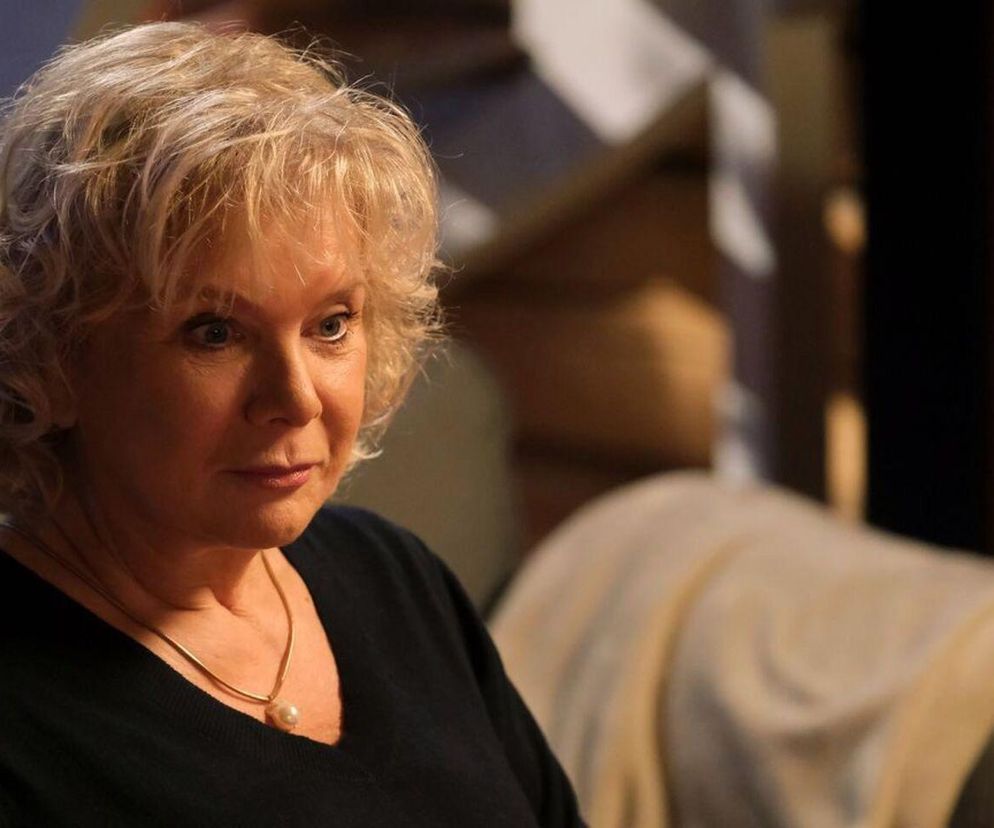 Aktorka Dorota Kamińska w roli Anny Lerman, matki Anity, zaniepokojona. Ma jasne, kręcone włosy, czarną bluzkę i naszyjnik z perłą. Zdjęcie oddaje napięcie związane z powrotem problemów z przeszłości, o czym przeczytasz więcej na Super Seriale.
