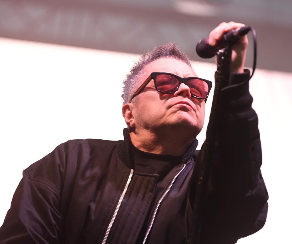 Muniek Staszczyk, lider zespołu T.Love, w czarnych okularach z czerwonawymi szkłami, trzymający mikrofon. Na zdjęciu widać jego głowę i część torsu w czarnej kurtce. Artykuł na Poradnik Zdrowie dotyczy choroby Staszczyka.