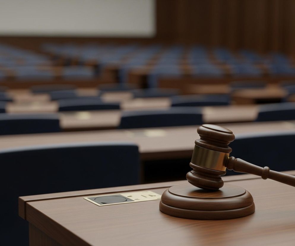 Młotek sędziowski wykonany z ciemnobrązowego drewna, z metalową obręczą w kolorze złota, leży na okrągłej podstawce z tego samego drewna, umieszczonej na drewnianym blacie stołu. Stół ten ma widoczną fakturę drewna w odcieniach brązu. Za nim rozciąga się rzędami pusta sala z kolejnymi drewnianymi stołami i ciemnymi, granatowymi krzesłami, które są rozmazane, co sugeruje płytką głębię ostrości. W tle widać jasną ścianę oraz fragmenty drewnianych paneli lub kurtyn w ciepłych odcieniach brązu, tworzących jednolity obraz.