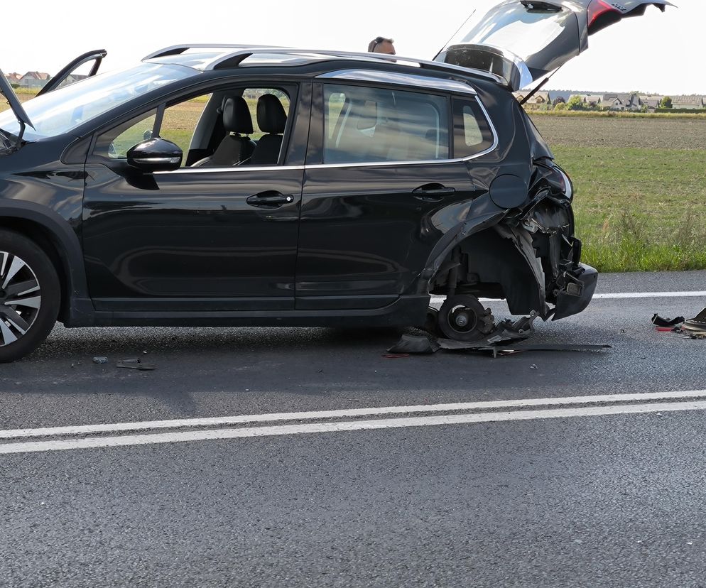 Czarny samochód z mocno uszkodzonym tyłem i otwartą maską stoi na poboczu drogi, z widocznymi odłamkami wokół tylnego koła, co ilustruje problem z uzyskiwaniem odszkodowań przez poszkodowanych, o czym czytasz na portalu Super Biznes.