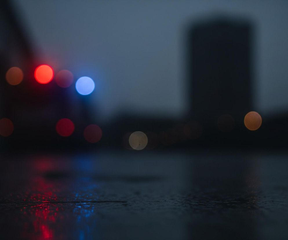 Na pierwszym planie widoczna jest mokra nawierzchnia, prawdopodobnie asfalt, pokryta drobnymi kroplami wody, odbijająca kolorowe światła. W tle, poza ostrością, widoczne są rozmyte punkty świetlne typu bokeh: duży, jasnoczerwony okrągły kształt po lewej stronie, obok niego mniejszy, ciemniejszy czerwony, a następnie jasnoniebieski okrąg. Dalej, po prawej stronie tła, znajduje się duża, rozmazana ciemna sylwetka budynku, a pod nią widoczne są mniejsze, rozmyte pomarańczowe i żółte światła. Całość utrzymana jest w ciemnych, chłodnych barwach, dominują odcienie granatu i szarości, podkreślając nocny lub bardzo pochmurny charakter sceny.