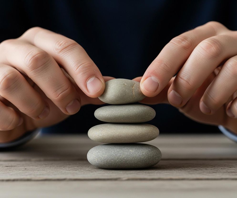 Męskie dłonie układają wieżę z pięciu gładkich, płaskich kamieni w odcieniach szarości i beżu. Dłonie są jasne, zadbane, z widocznymi paznokciami, a ich palce wskazujące i kciuki delikatnie trzymają górny kamień. Ubrana w ciemnoniebieski sweter lub marynarkę osoba opiera dłonie na szorstkim, drewnianym blacie stołu, którego faktura jest wyraźnie widoczna. Tło jest rozmyte i ciemne, w jednolitym odcieniu granatu.